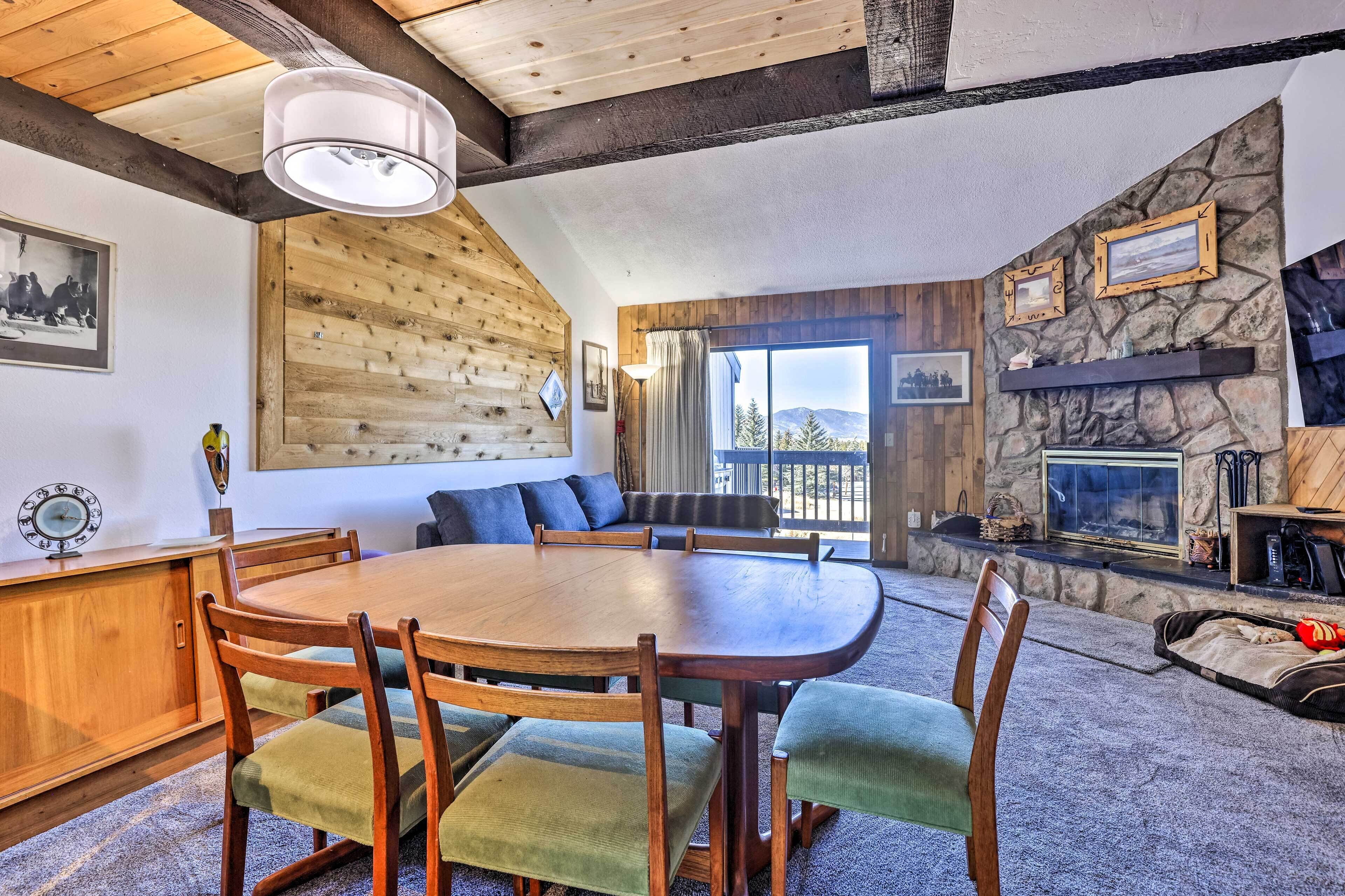 A cozy living room with a wooden dining table, comfortable seating, a stone fireplace, and a large window offering a view of the outdoors.