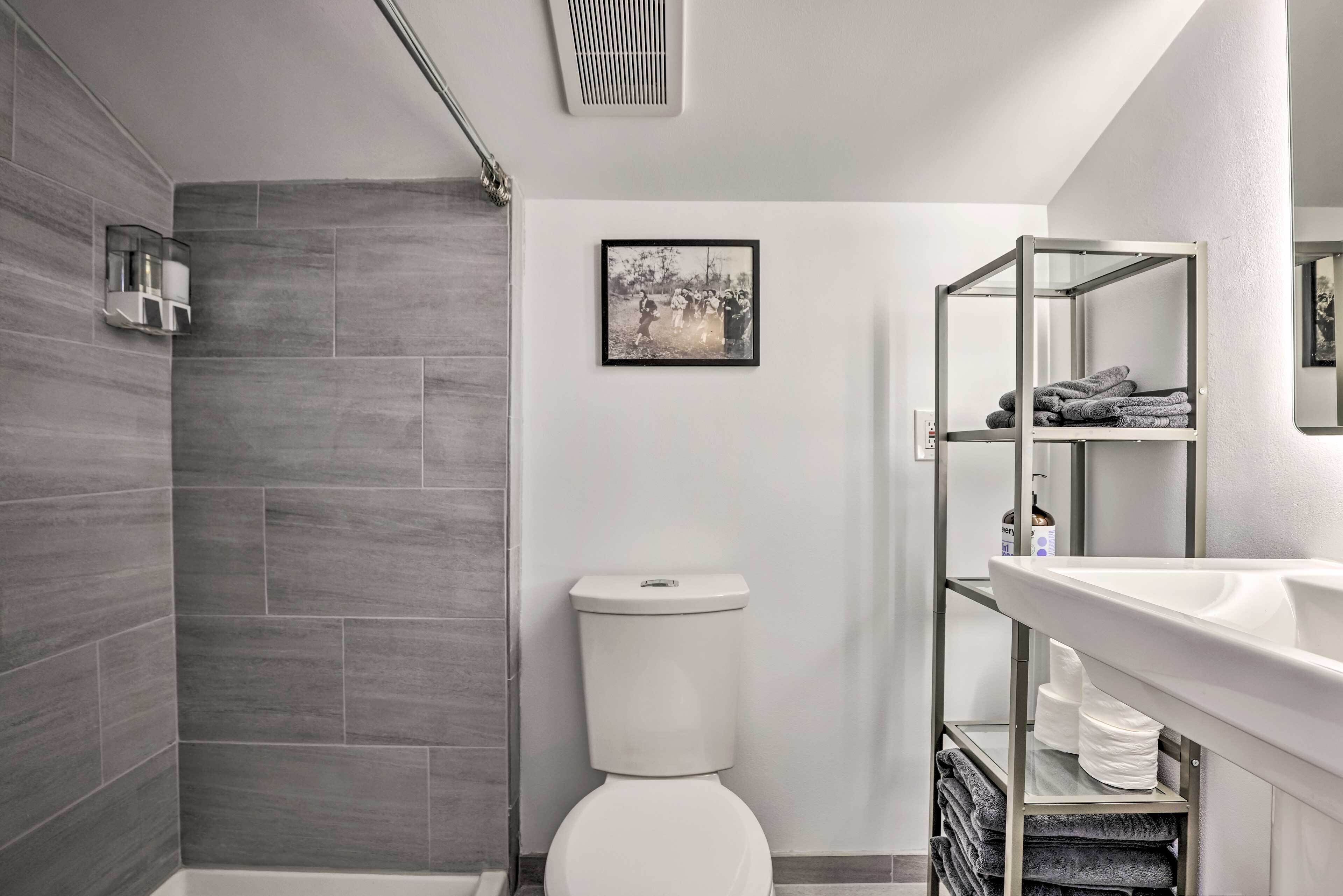 A modern bathroom with grey tiles, a white toilet, a shower, a sink with a mirror, and a towel rack.