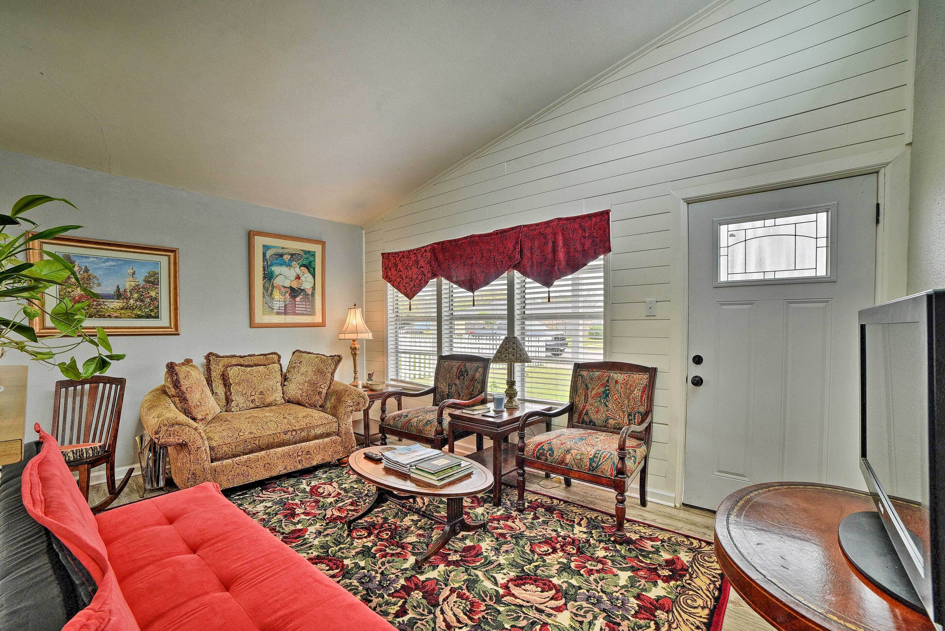 Un salon avec un tapis à motifs, un canapé rouge, une table basse en bois et deux fauteuils. On y trouve également un téléviseur posé sur un meuble en bois, une porte avec une fenêtre et un tableau accroché au mur.