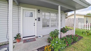 A house with a white front door, a covered porch, and a brick flower bed with various plants and flowers.