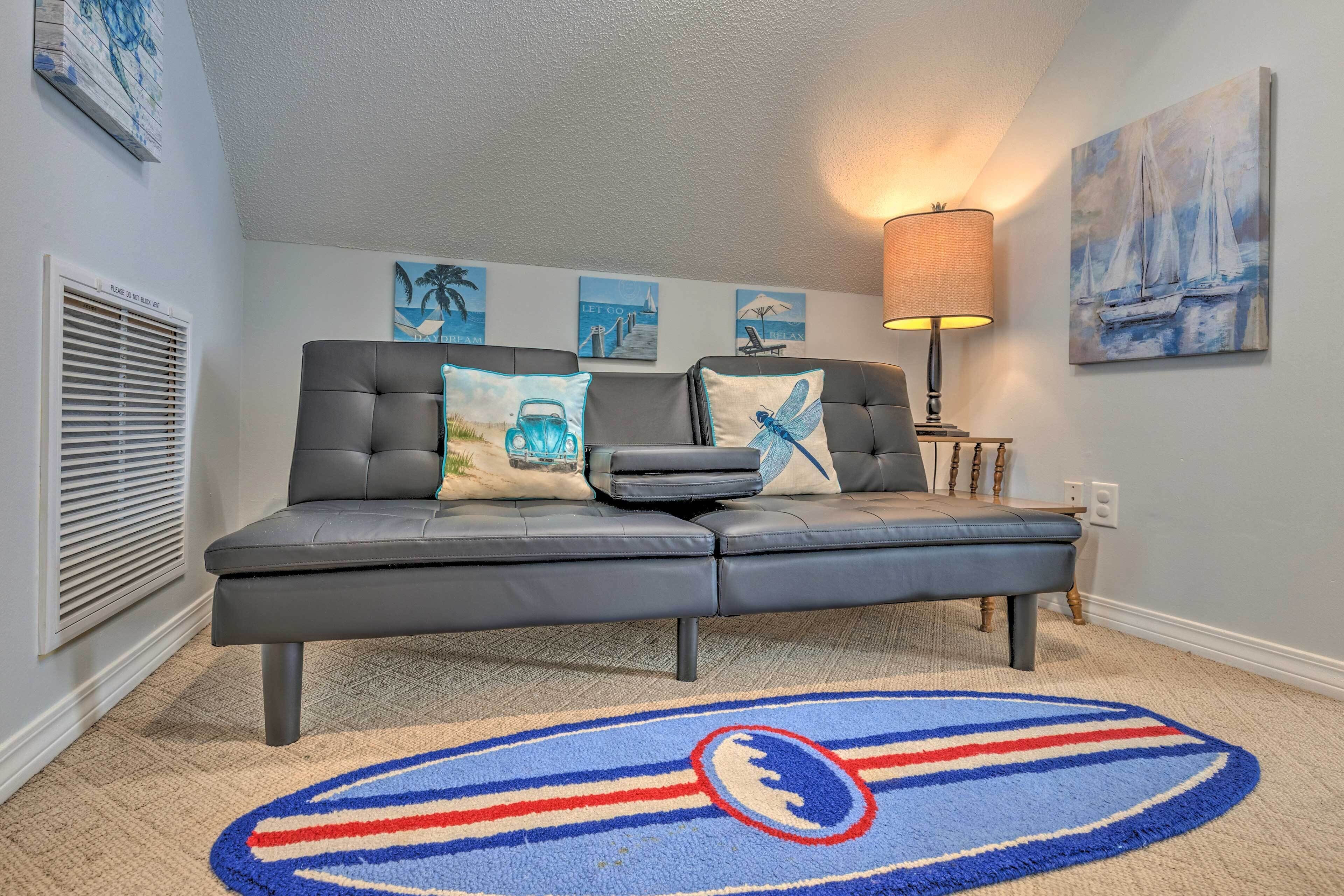 Ein modernes Wohnzimmer mit einem grauen Ecksofa, einem blauen und roten Streifen Teppich sowie Wandbildern von Segelbooten und Palmen.