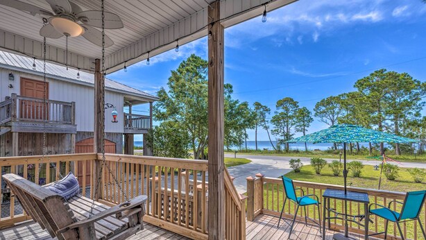 A wooden deck with a swing chair, a table with chairs, and a view of the beach and trees. - Stunning Views & Waterfront Fun: Aloe Bay Home (Dauphin Island)