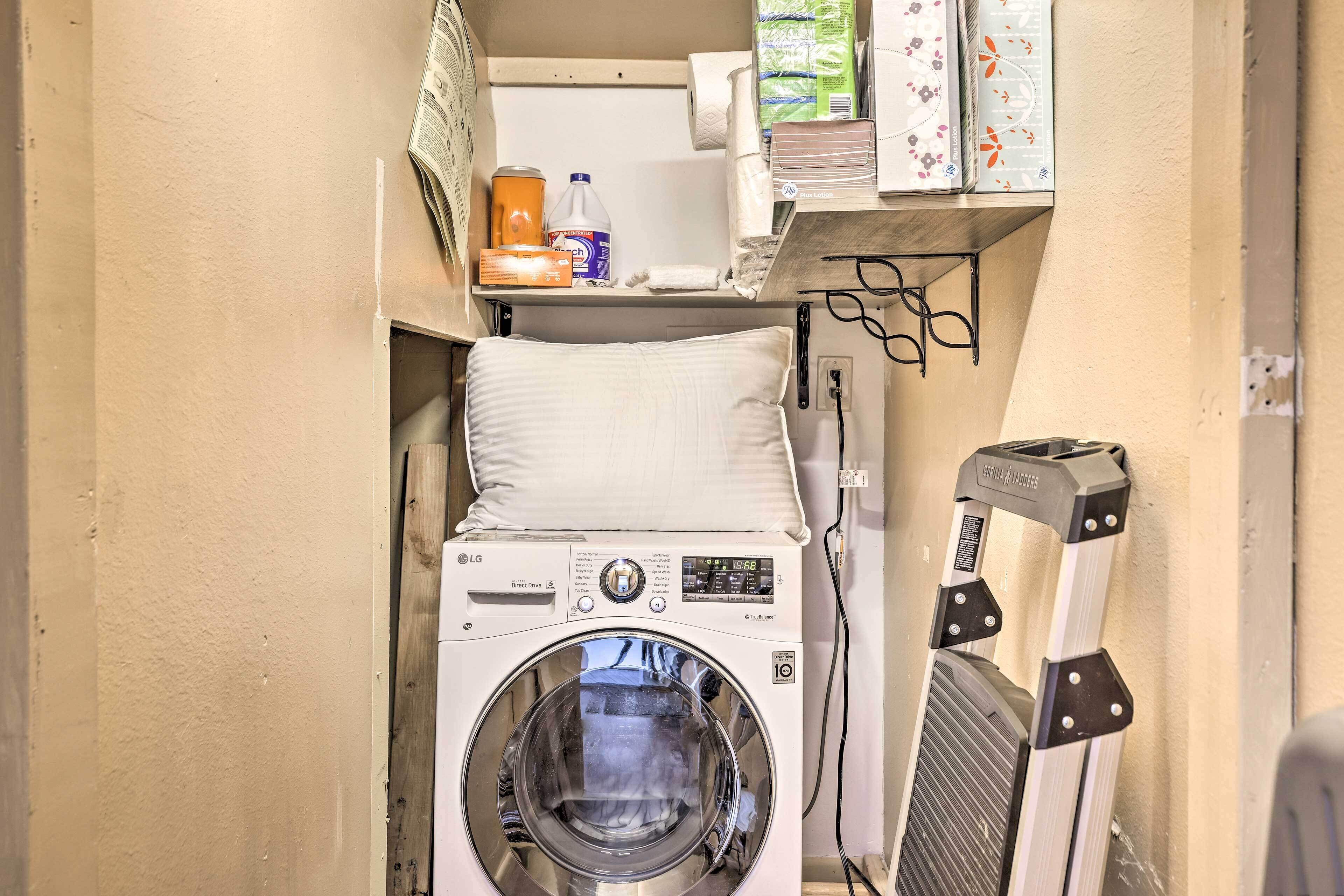 A compact laundry area with a front-loading washing machine, a drying rack, and a shelf with cleaning supplies.