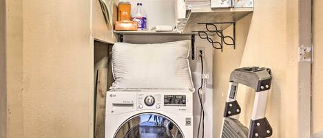 A compact laundry area with a front-loading washing machine, a drying rack, and a shelf with cleaning supplies.