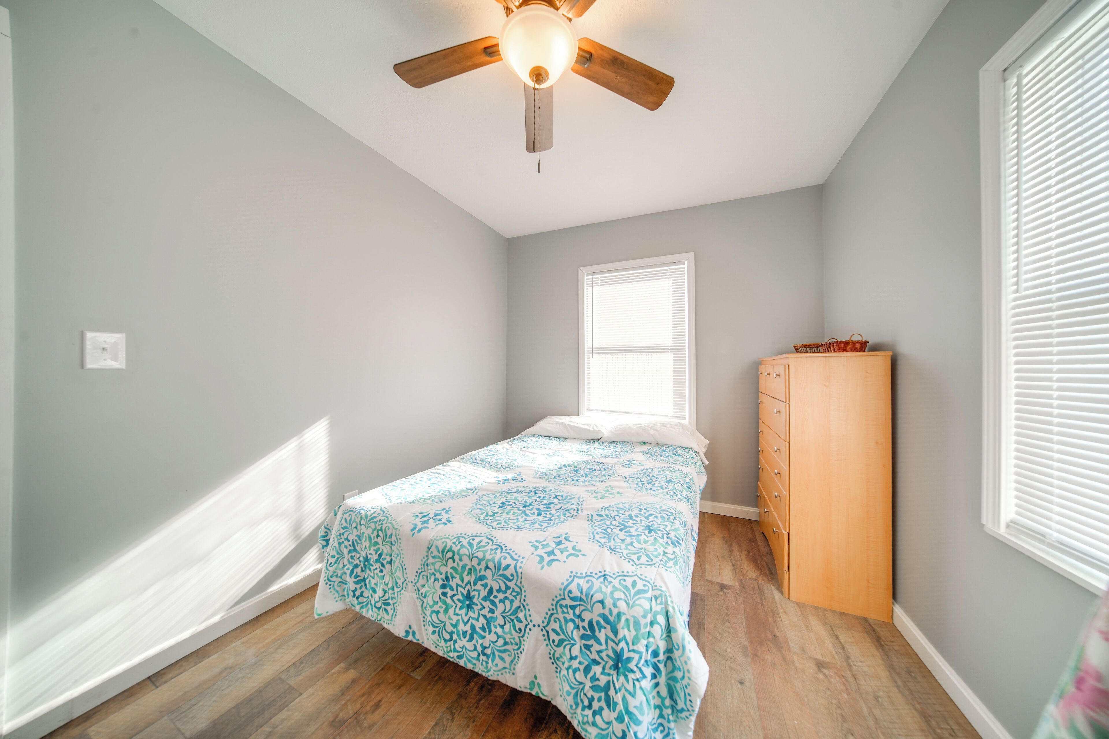 A bedroom with a bed, a wooden dresser, a ceiling fan, and a window with blinds.