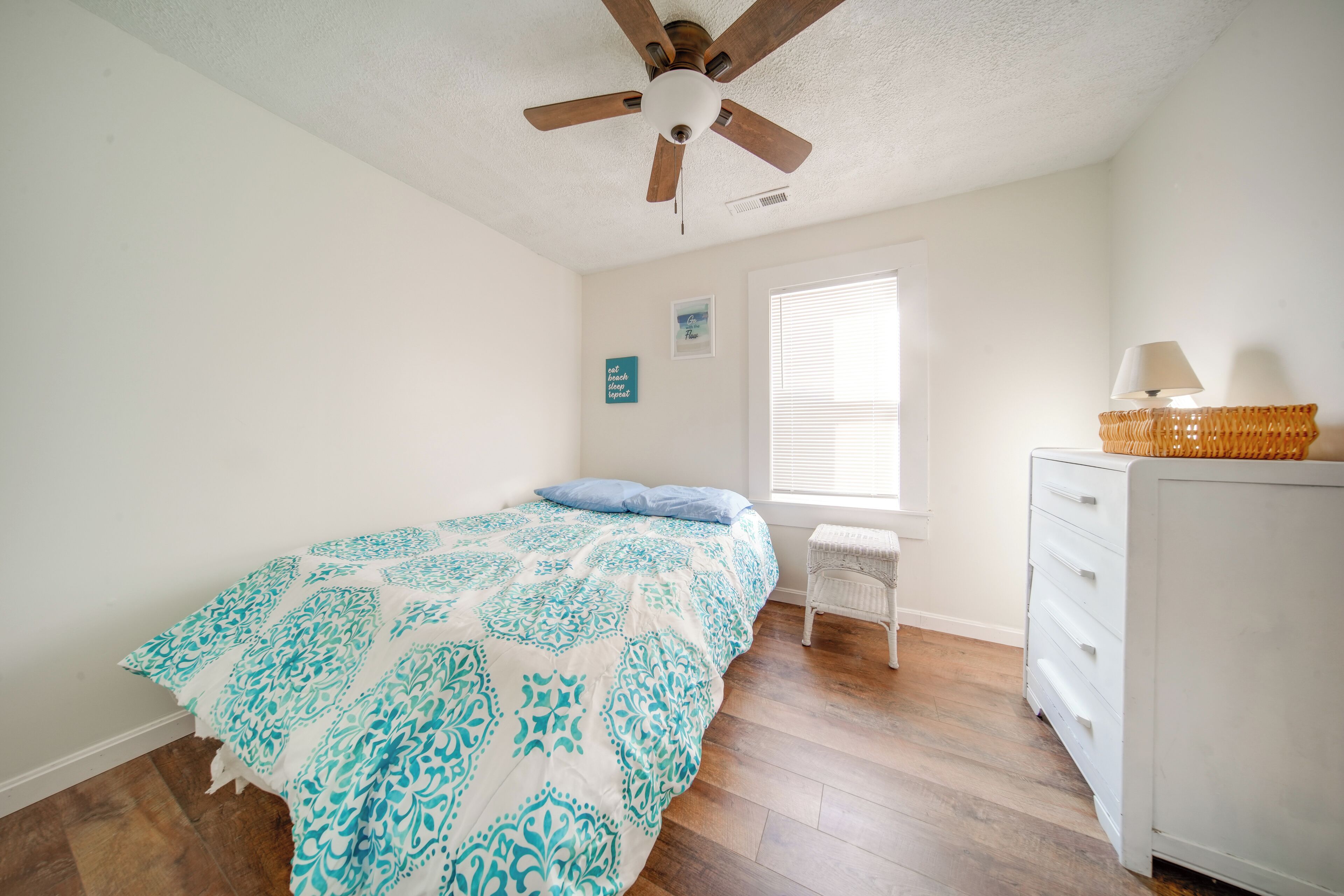 Un dormitorio con cama, ventilador de techo, ventana con persianas, cómoda blanca y una mesita.