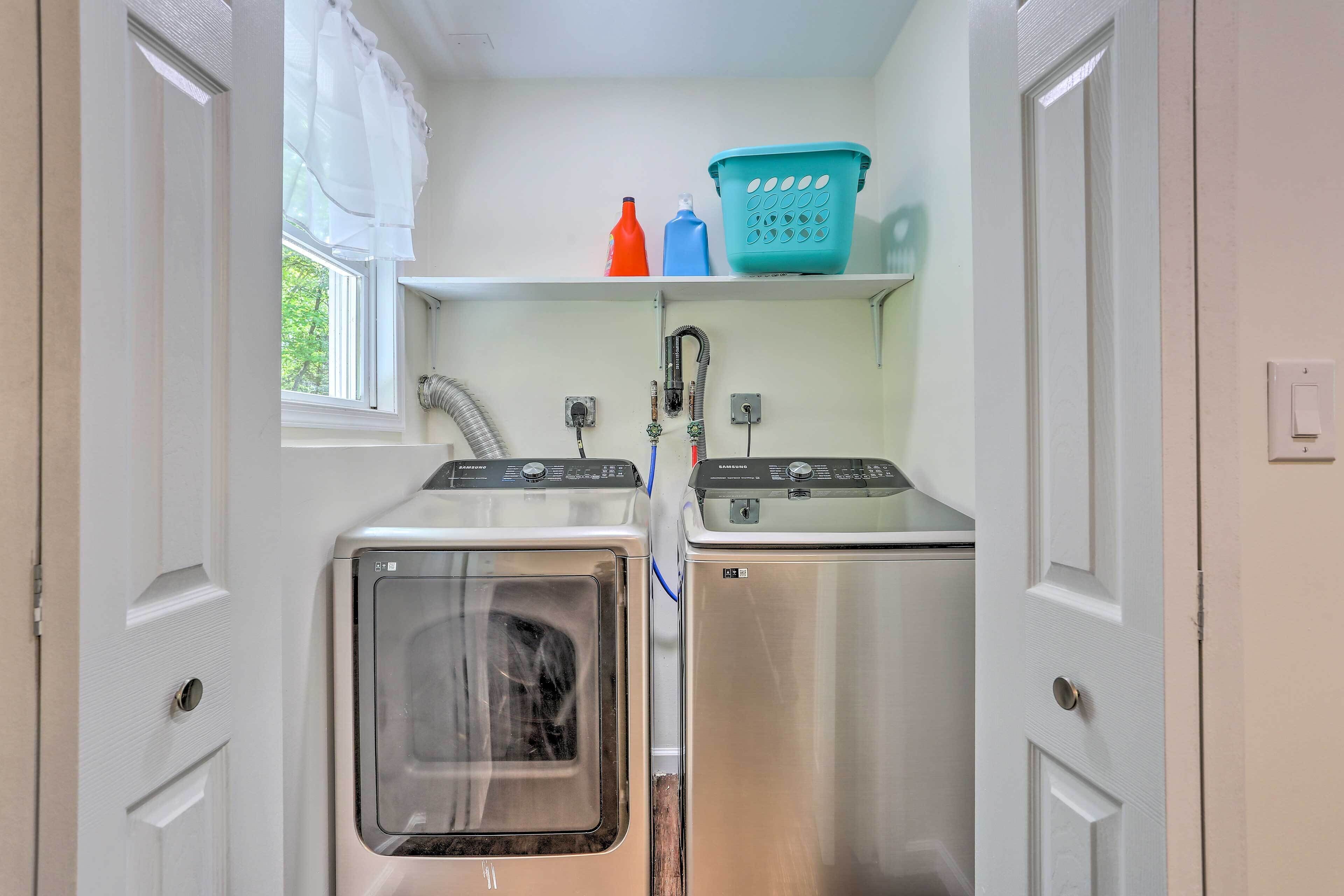 A compact laundry room with two stainless steel washers and dryers, a window with white curtains, and a shelf with laundry supplies.