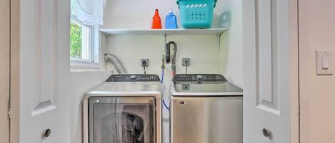 A compact laundry room with two stainless steel washers and dryers, a window with white curtains, and a shelf with laundry supplies.