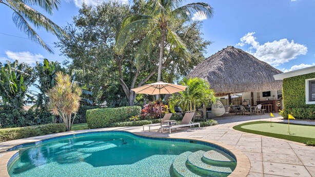 A swimming pool with a thatched roof structure, surrounded by palm trees and a well-maintained lawn. - Poolside Paradise w/ Tropical Hut in Palm Beach (Palm Beach Gardens)