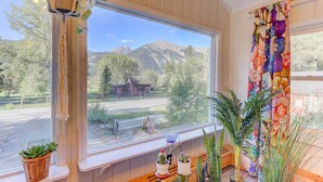 A room with a large window overlooking a mountain and a road, with a white picket fence and a house in the background. - Remodeled Lake Cabin, Stunning Mtn View, New Patio (Twin Lakes)