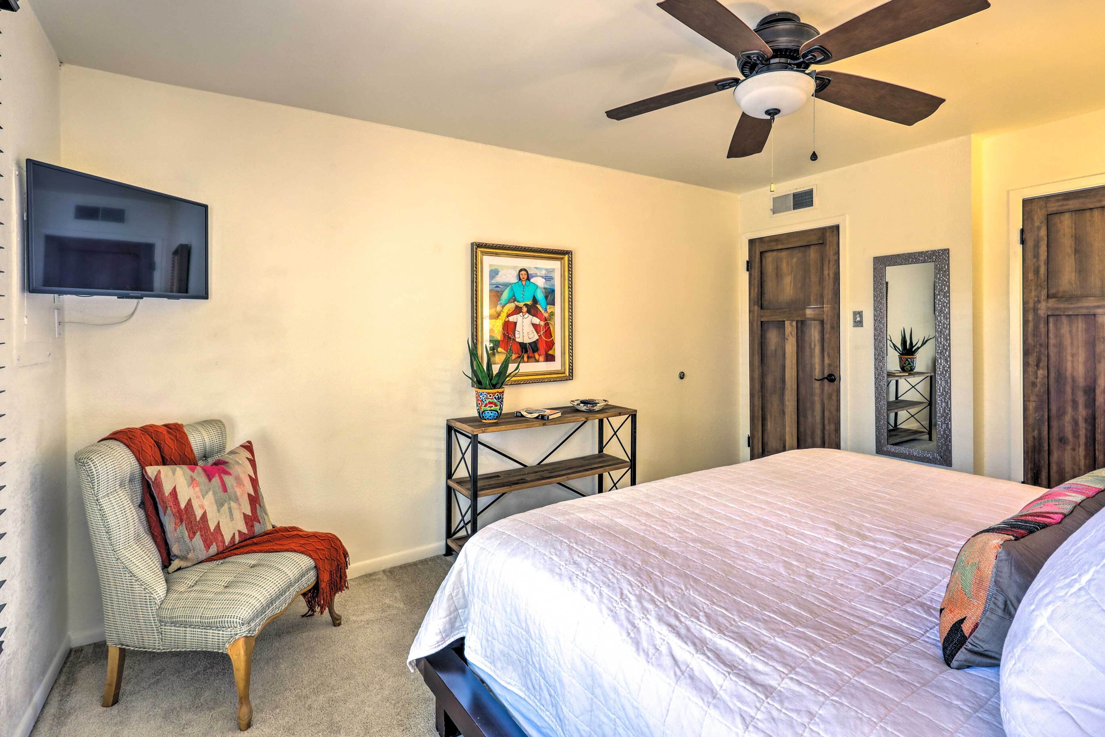 A bedroom with a bed, a chair, a TV mounted on the wall, a ceiling fan, and a framed picture on the wall.