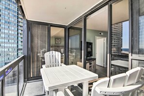 A balcony with a white table and chairs, a sliding glass door leading to a kitchen area, and a view of a cityscape.