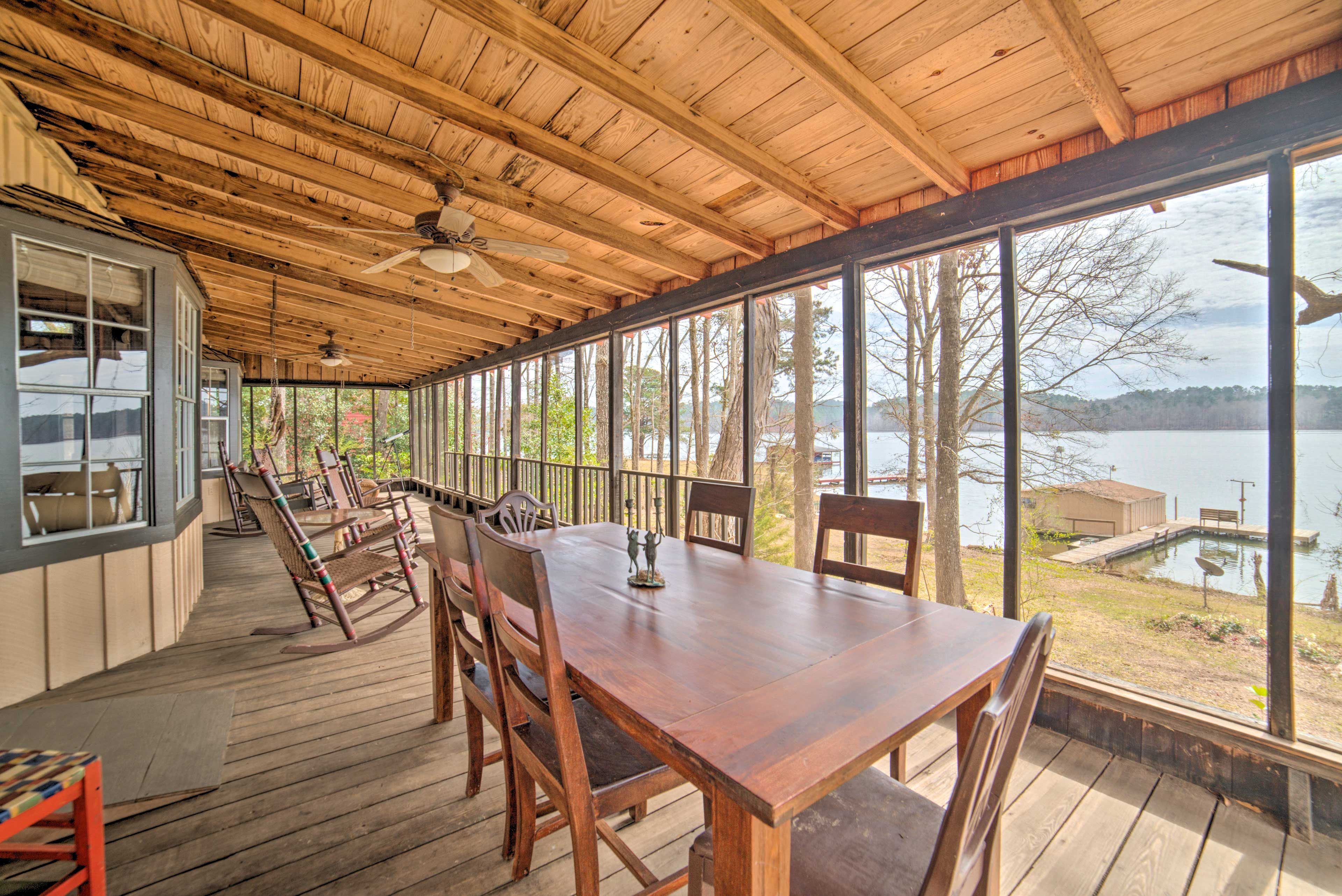 A covered outdoor deck with a wooden table and chairs, large windows offering a view of a lake and dock.