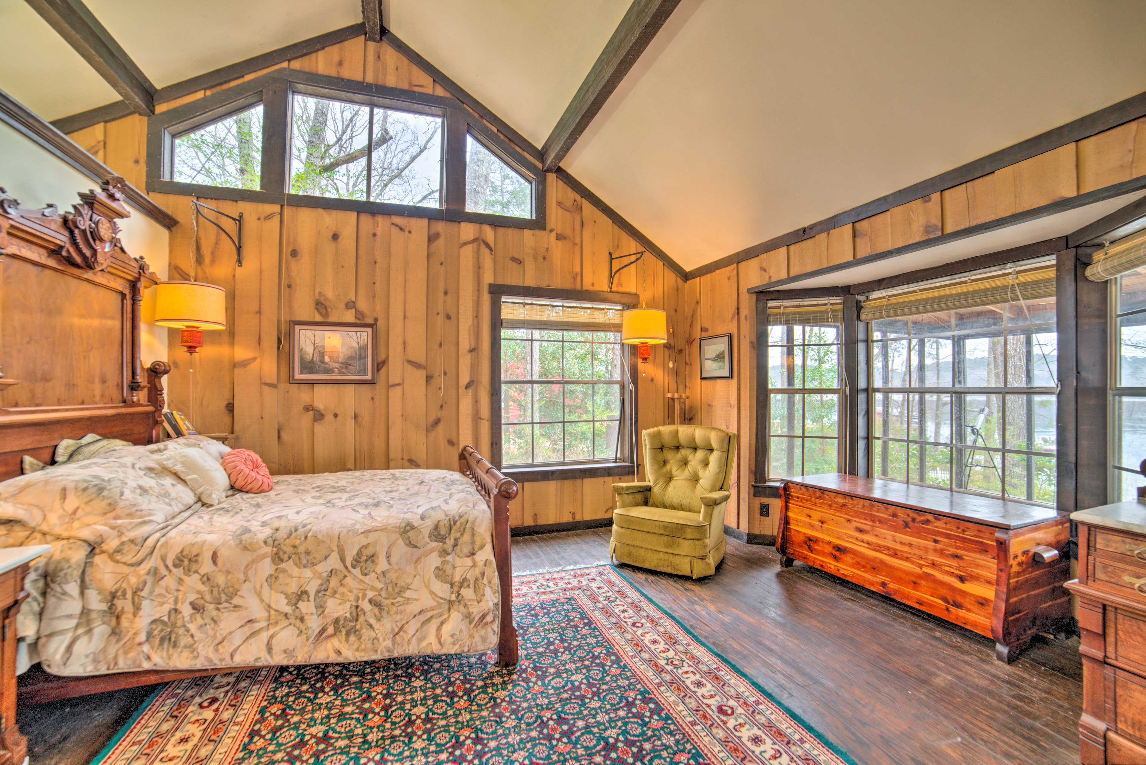 A spacious bedroom with a large bed, a chair, a dresser, and multiple windows offering natural light and views.