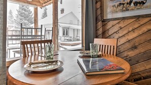 A wooden dining table with a book and glasses, a framed picture on the wall, and a view of a house through a window. - Cozy Log Cabin Escape In the Heart of Creede! (Creede)