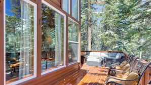 Una terraza de madera con mobiliario exterior y amplios ventanales con vistas a un área boscosa.