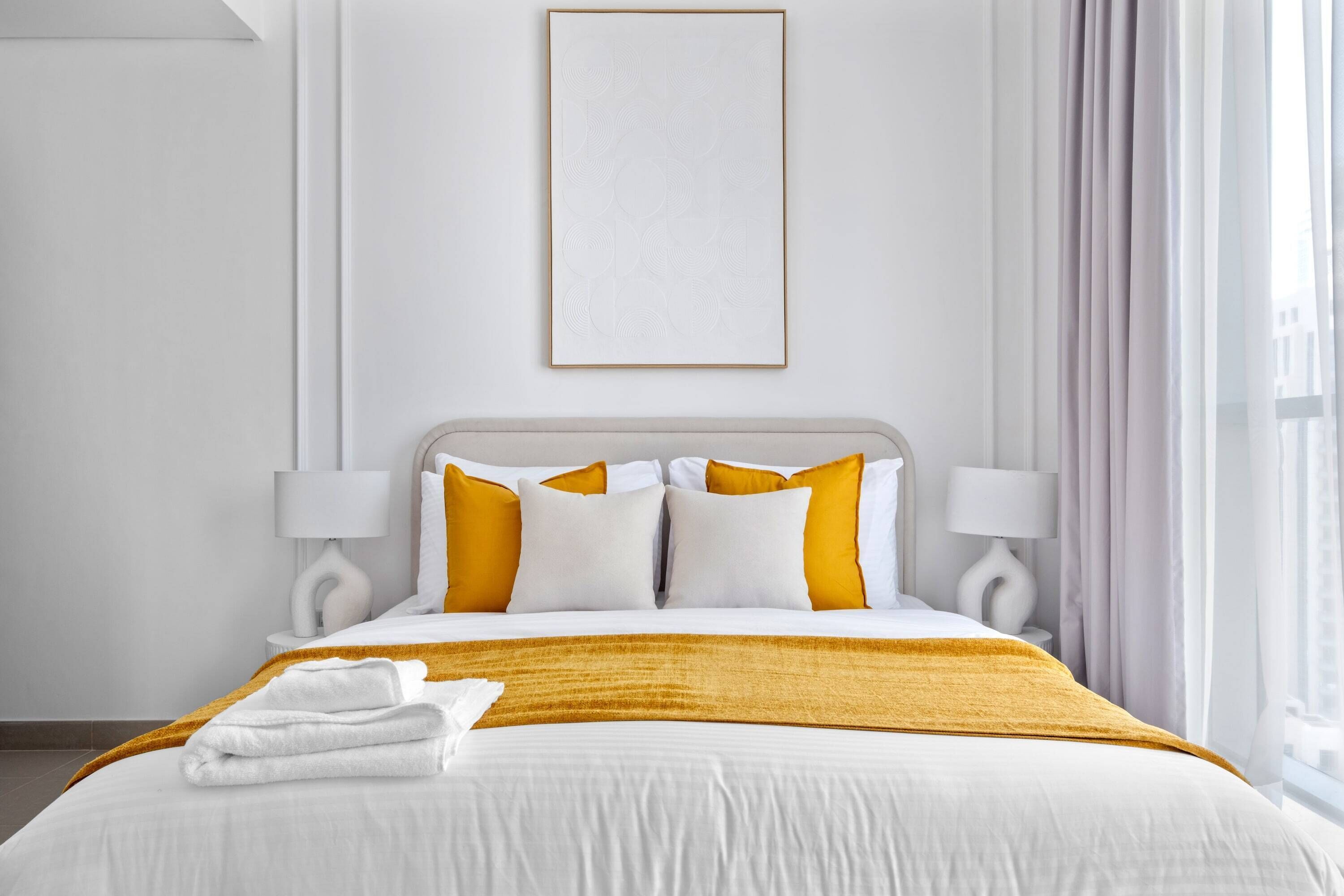 Ein Hotelzimmer mit einem großen Bett, weißer Bettwäsche und einer senfgelben Zudecke. Zwei Nachttischlampen und ein gerahmtes Bild sind an der Wand zu sehen.