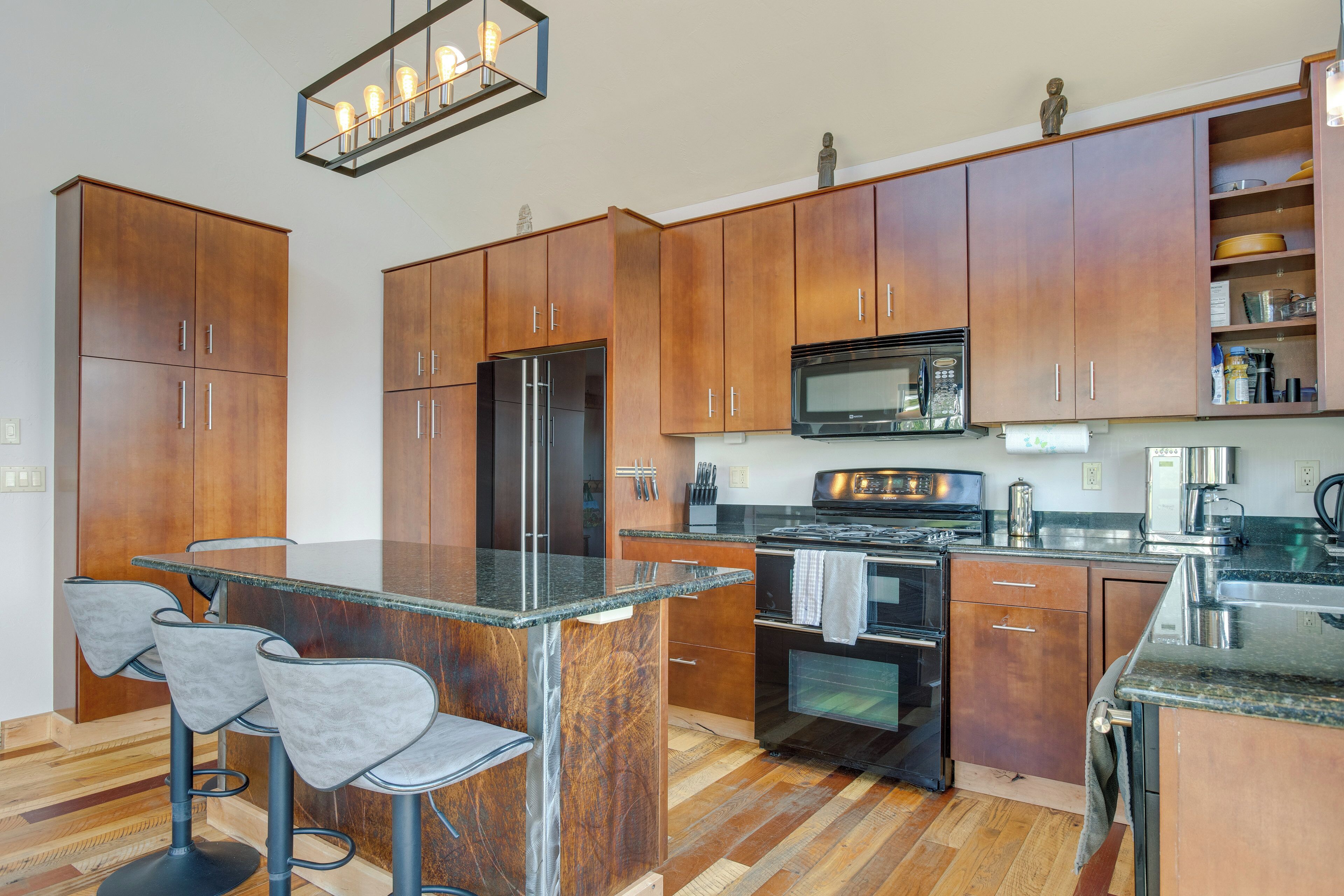 A modern kitchen with wooden cabinets, a stainless steel refrigerator, a microwave, and a stove. There are three bar stools and a granite countertop.