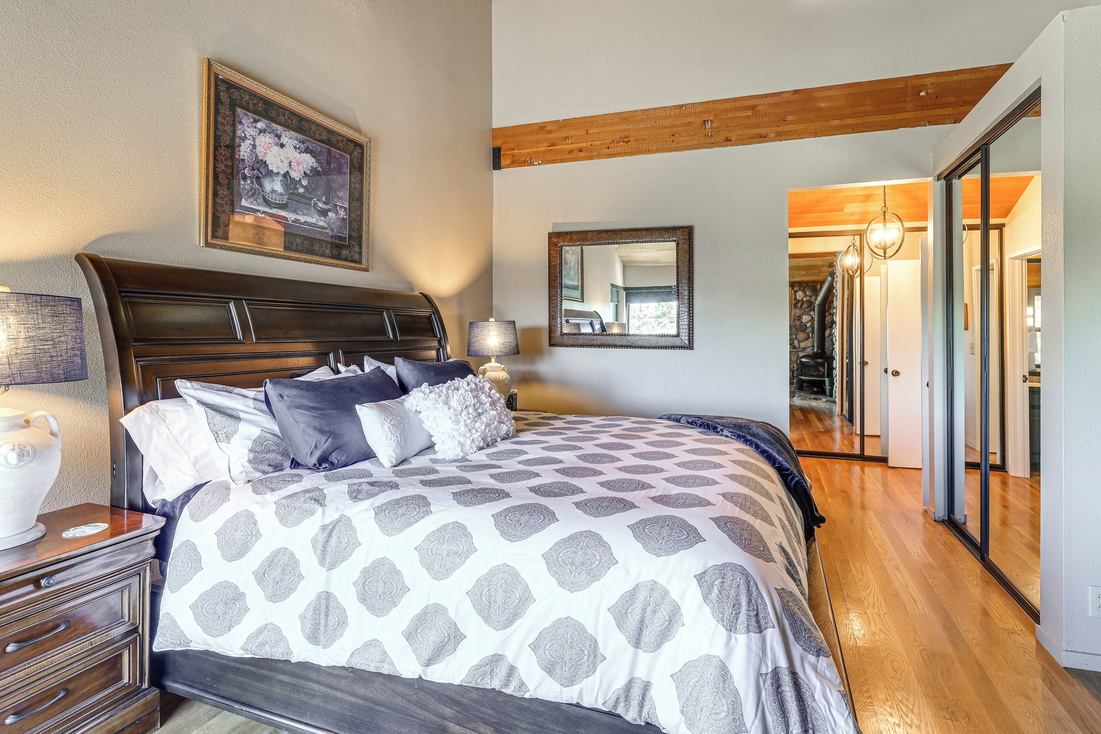 A bedroom with a large bed, wooden headboard, a nightstand with a lamp, a framed picture on the wall, and a hallway leading to another room.