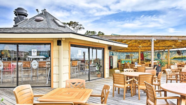 Un patio al aire libre con mesas y sillas de madera, un espacio de estar cubierto y vistas de la ciudad a través de grandes ventanales.