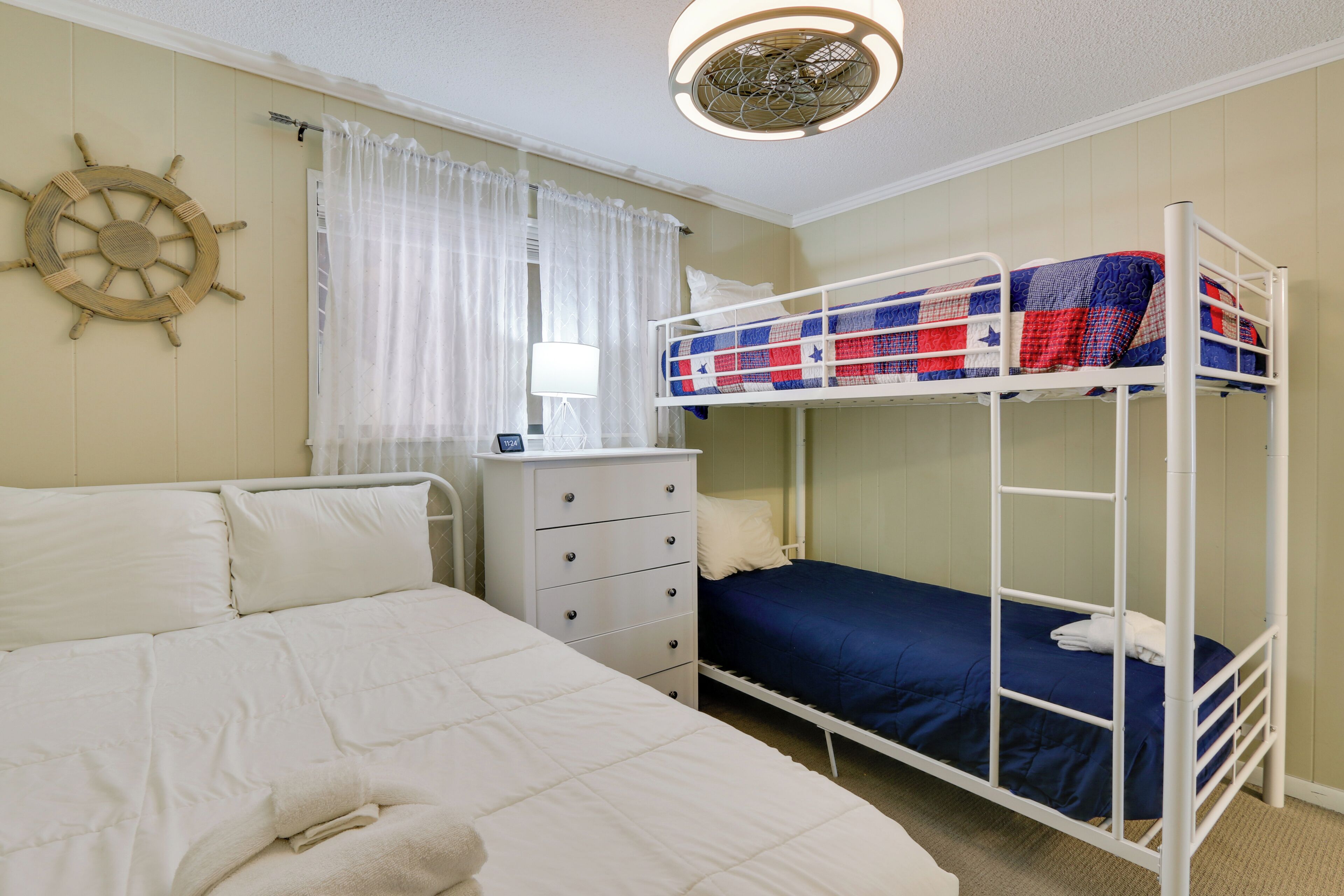 Une chambre avec un lit superposé, un lit blanc, une commode, un décor représentant un gouvernail de bateau et une fenêtre avec des rideaux.