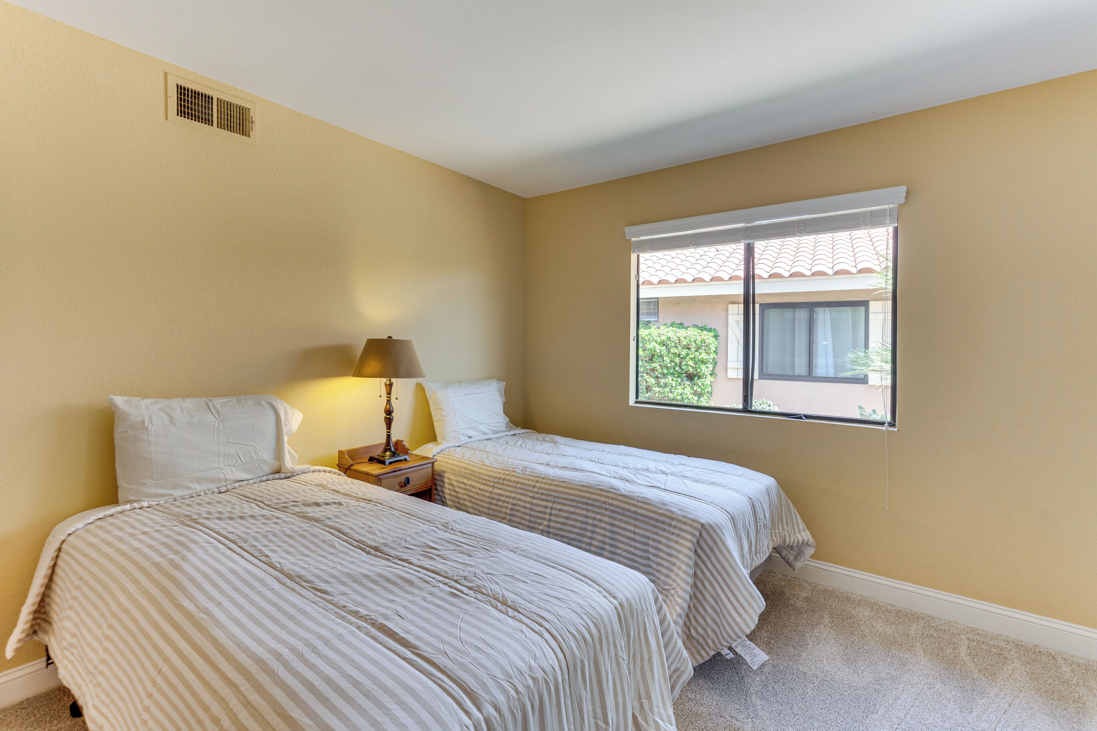 A bedroom with two beds, a nightstand, a lamp, and a window with a view of greenery.