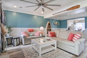 A living room with a ceiling fan, a glass coffee table, two sofas with striped pillows, a small side table, and a decorative shelf. - Walk to Maryland Beach: Canal-front Escape w/ Dock (Ocean City)