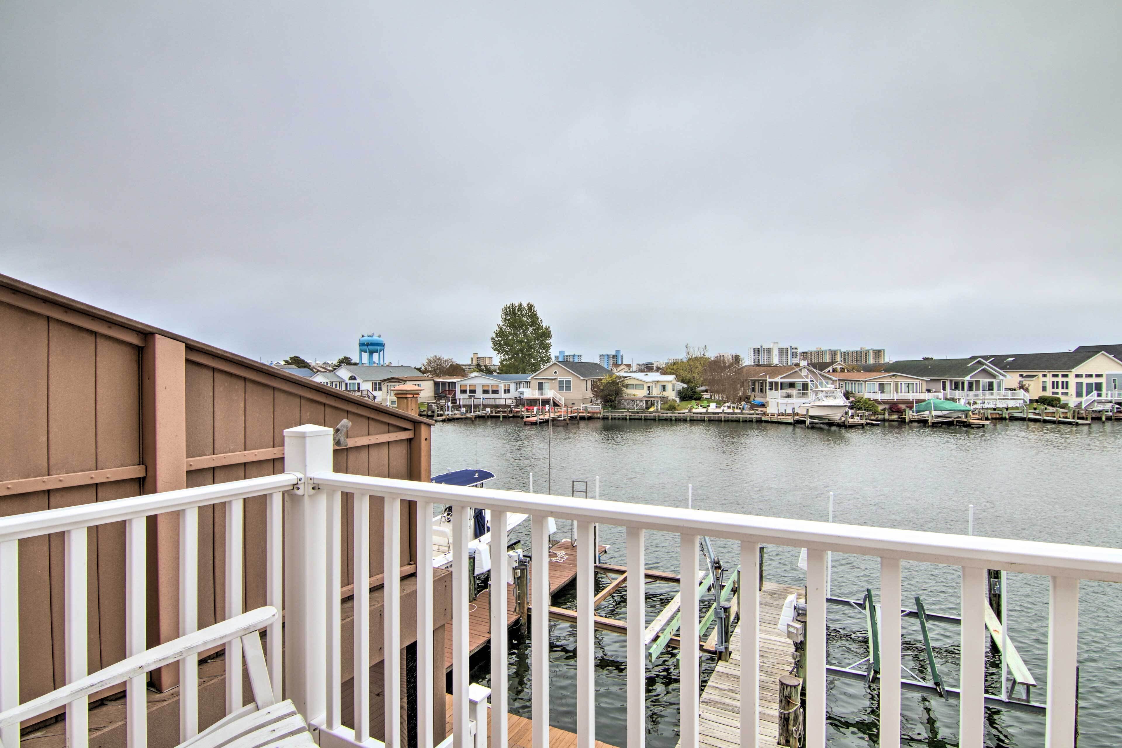 A waterfront view from a balcony with a wooden deck, docked boats, and a row of houses across the water.