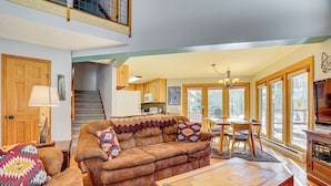 A spacious living room with a brown leather sofa, a wooden coffee table, and a dining area with a table and chairs. A staircase leads to an upper level, and large windows provide natural light. - 10 Mi to Keystone Resort: Cabin w/ Private Hot Tub (Silverthorne)