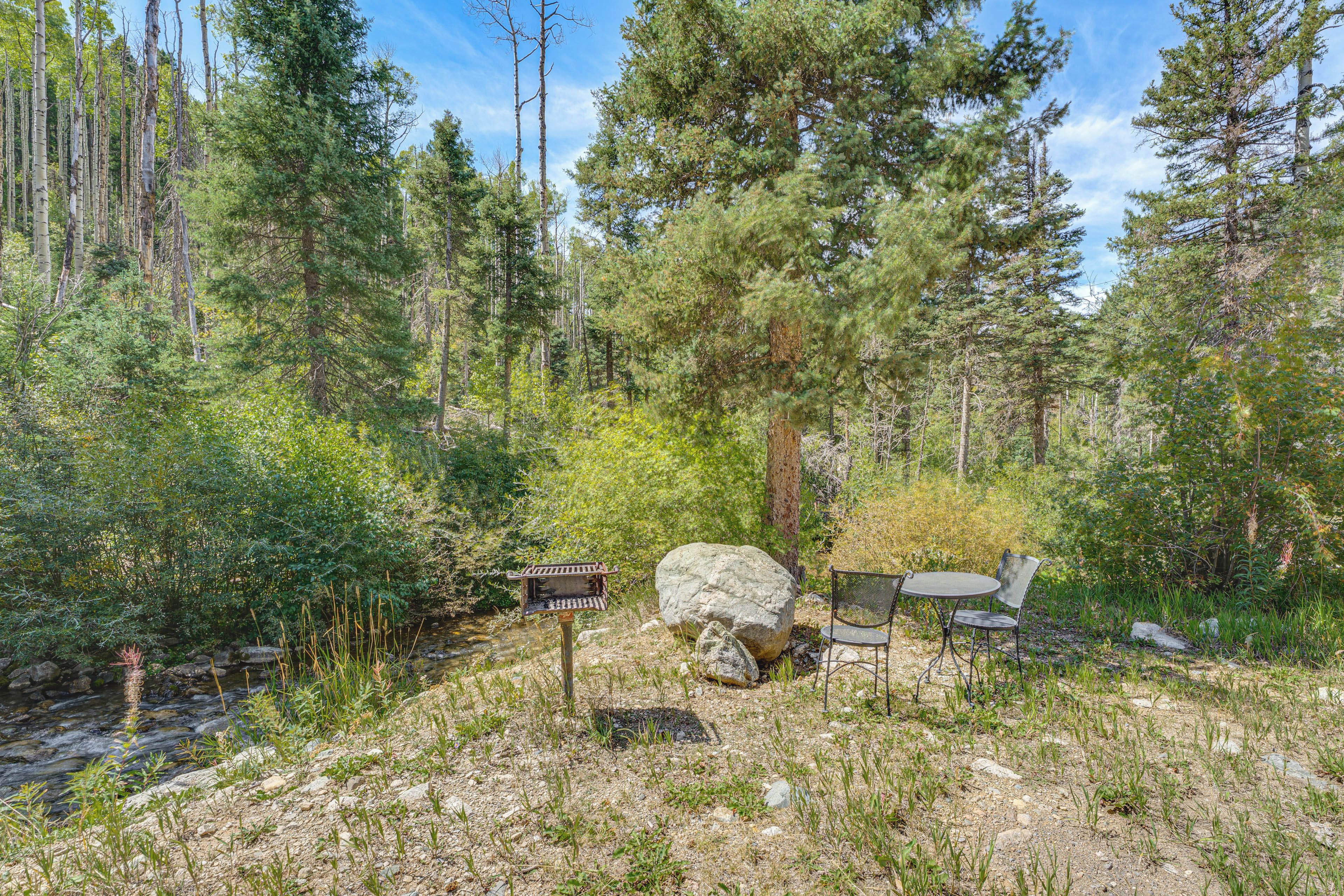 Eine natürliche Umgebung im Freien mit einem Picknicktisch und -stühlen, umgeben von Bäumen und einem kleinen Bach.