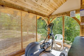Ein überdachter Fitnessbereich im Freien mit Trainingsgeräten, darunter ein Ergometer und ein Laufband, sowie einem Stuhl.