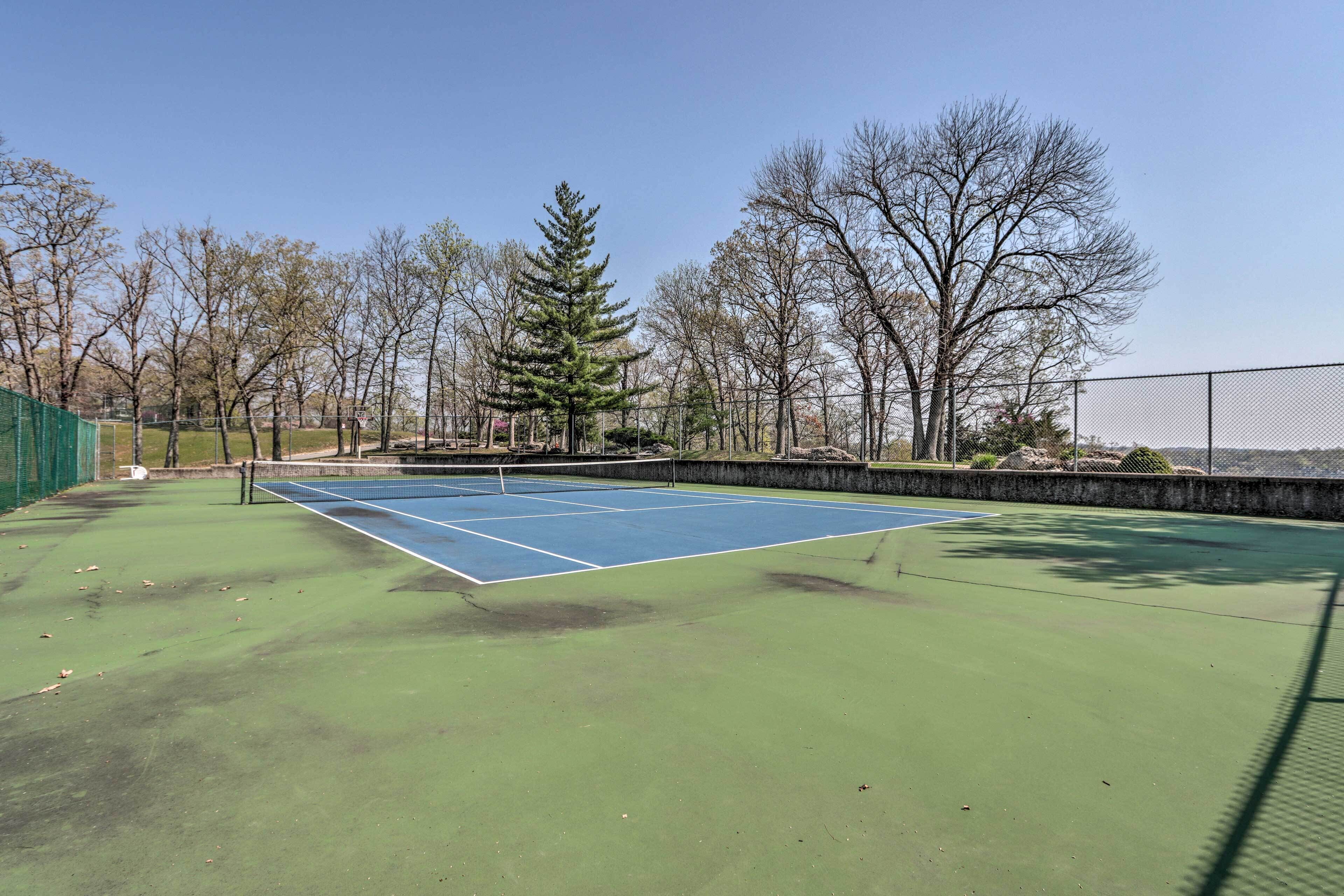 An outdoor tennis court with a green surface, surrounded by trees and a fence.