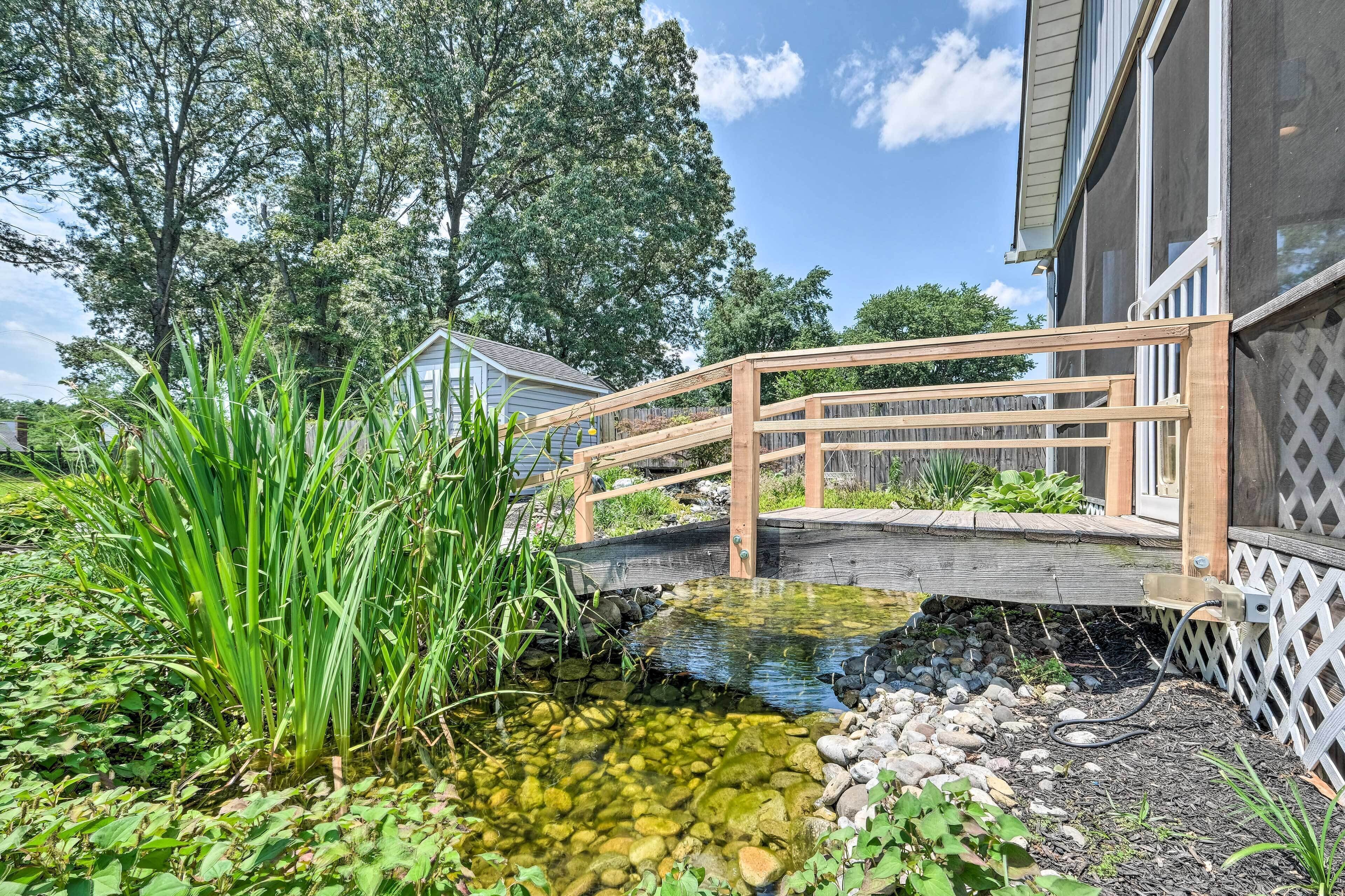 Un puente de madera sobre un estanque cristalino con rocas y plantas, rodeado de árboles y una casa.