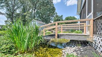 Un puente de madera sobre un estanque cristalino con rocas y plantas, rodeado de árboles y una casa.