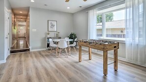 A spacious room with a wooden floor, a ceiling fan, a foosball table, a dining area with chairs and a table, and a large window with curtains. - Idyllic Pasadena Retreat With Garden + Hot Tub! (Pasadena)