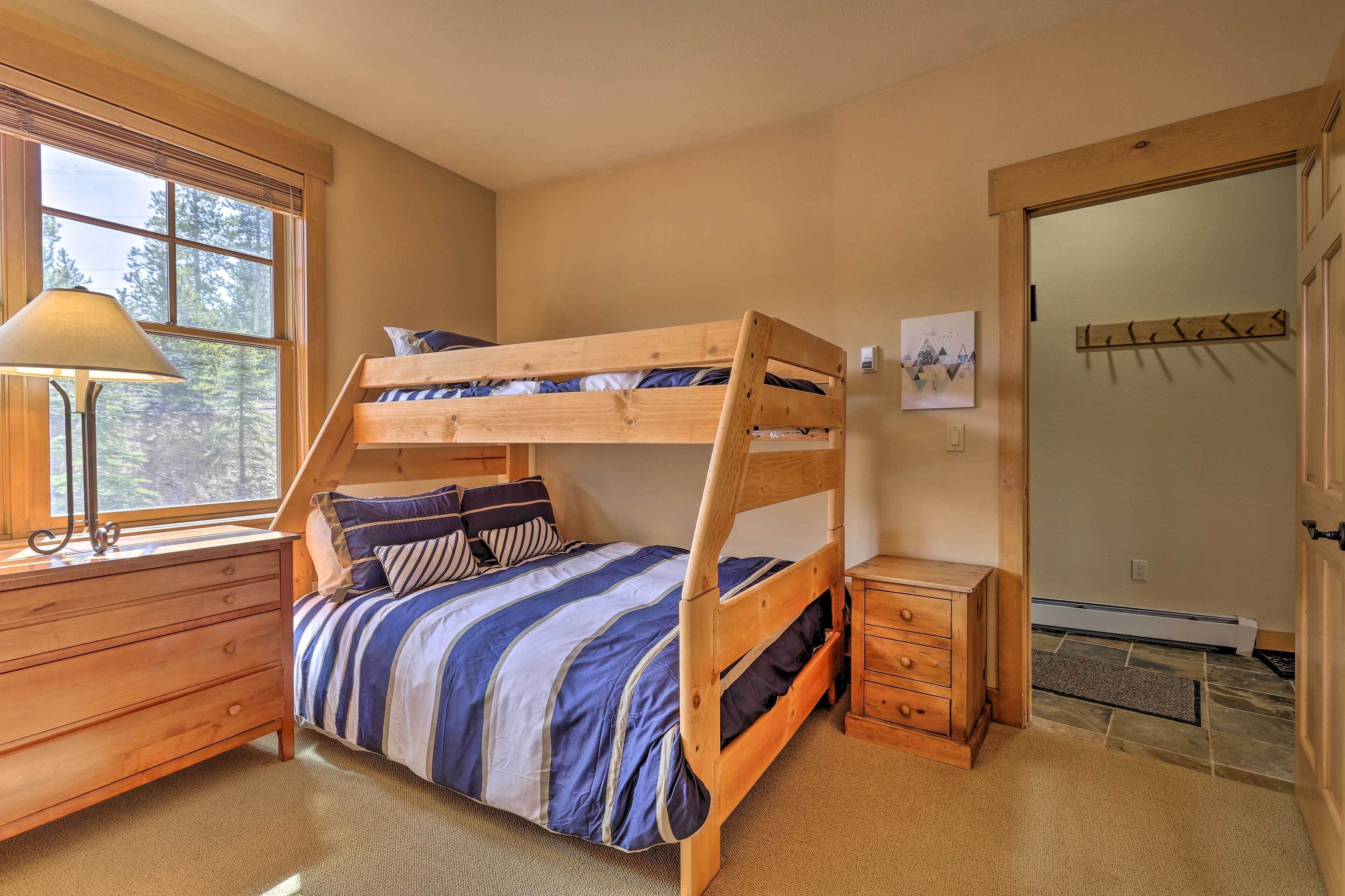 Un lit superposé avec une couvre-lit à rayures, une commode en bois, une lampe et une fenêtre donnant sur les arbres.