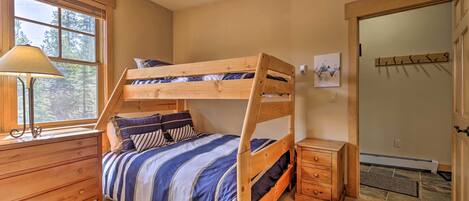 Un lit superposé avec une couvre-lit à rayures, une commode en bois, une lampe et une fenêtre donnant sur les arbres.