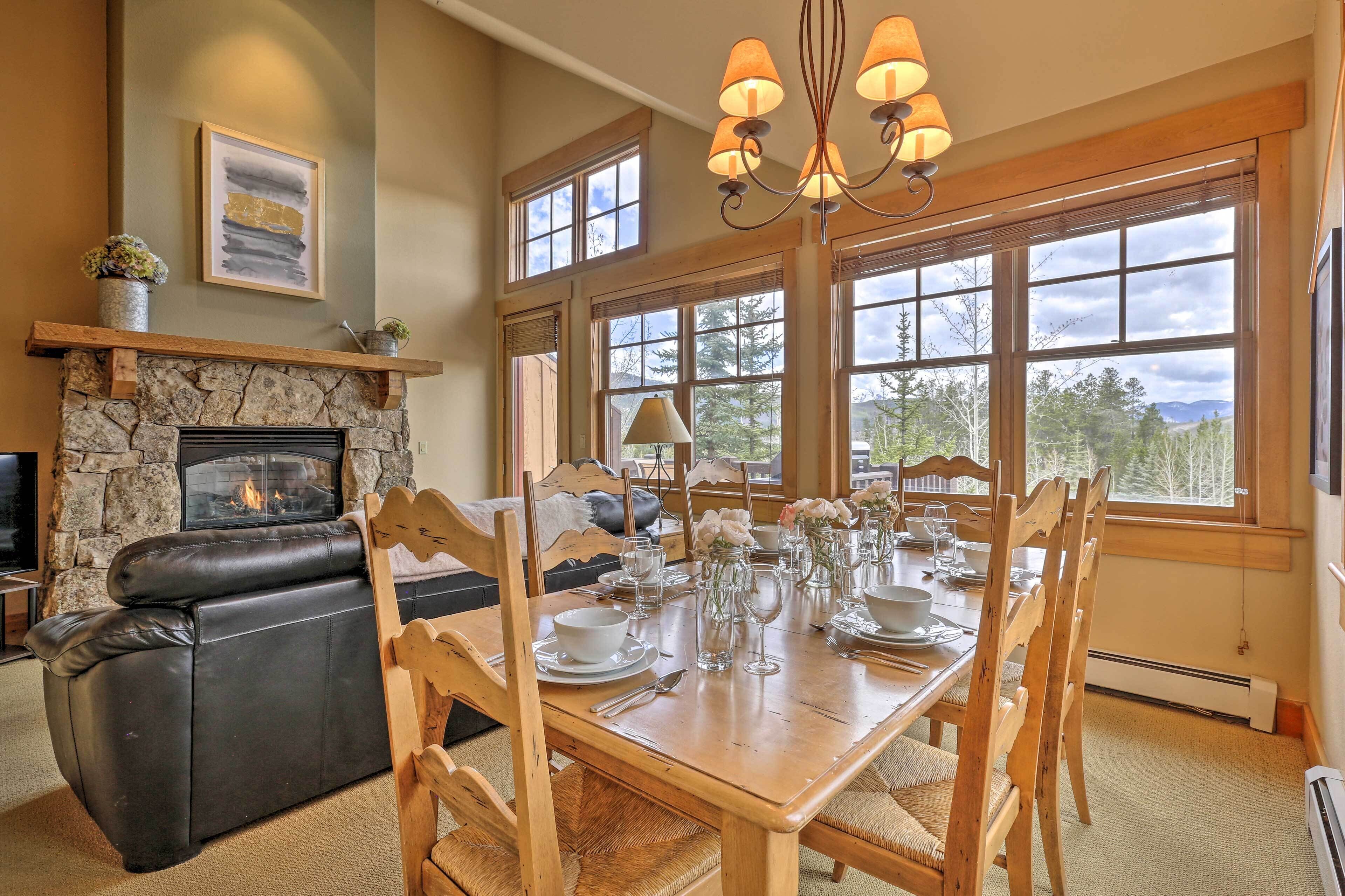 A cozy living room with a fireplace, a dining table set for four, and a large window offering a view of trees and mountains.