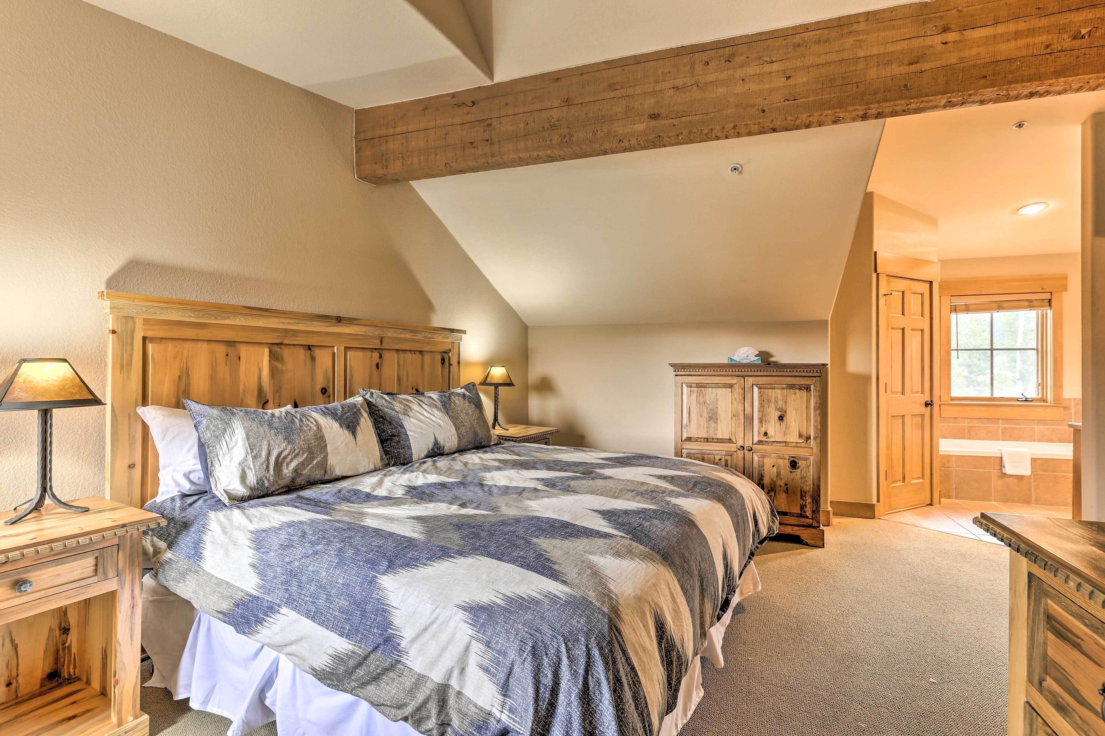 Quarto com uma cama grande, cabeceira de madeira, mesa de cabeceira com abajur, cômoda e porta para outro espaço.