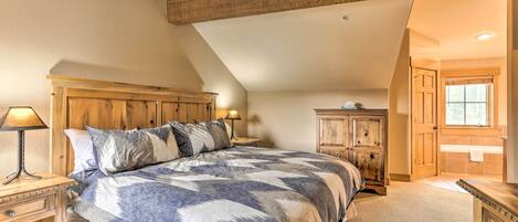 Quarto com uma cama grande, cabeceira de madeira, mesa de cabeceira com abajur, cômoda e porta para outro espaço.