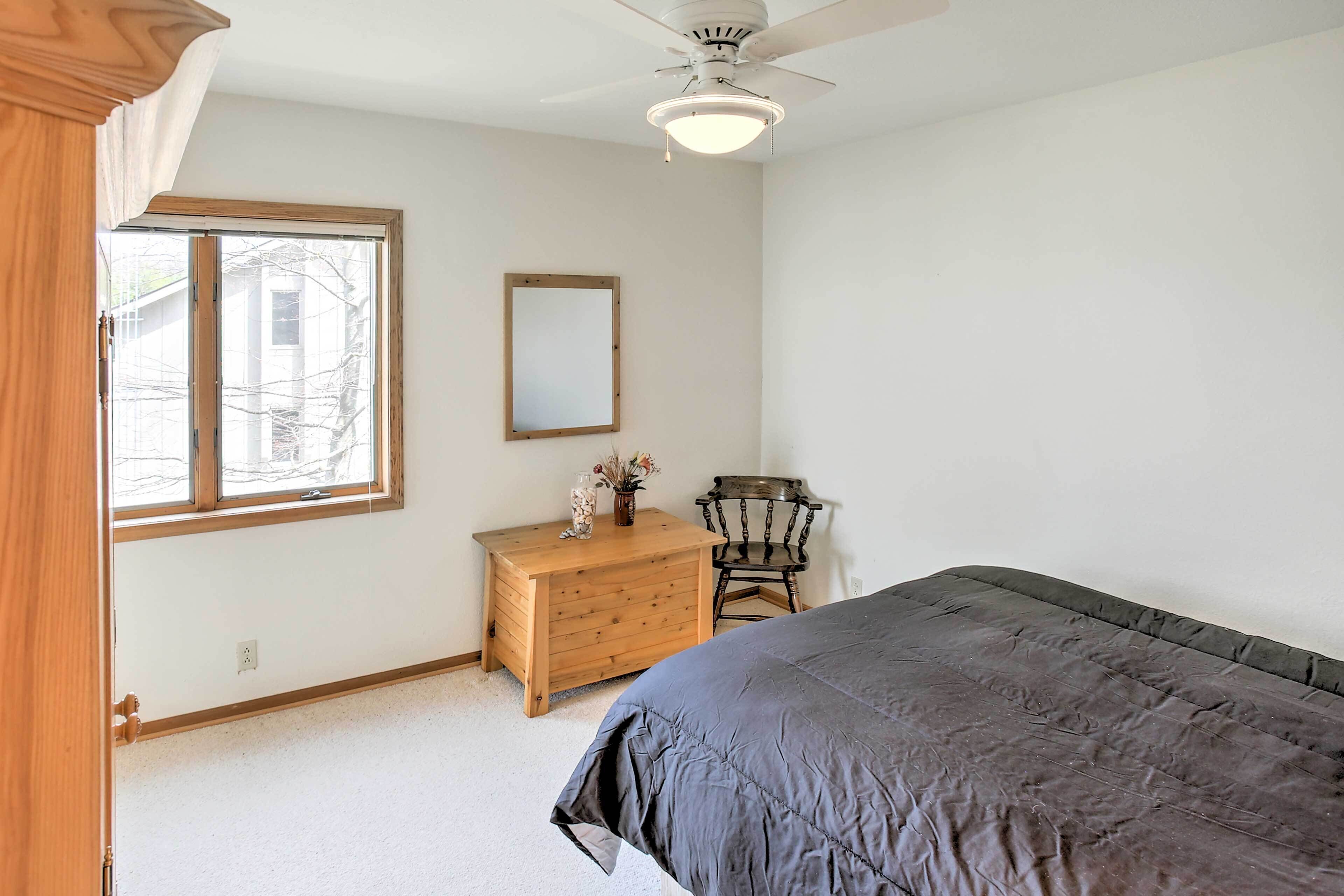 Quarto com cama, cômoda de madeira, cadeira, ventilador de teto e janela.