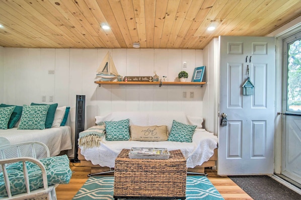 A cozy room with a wooden ceiling, a white sofa with patterned cushions, a small table with a tray, and a door with a hanging ornament. - Walk to Beach: Bright & Cozy Cape Cod Studio! (Dennis Port)