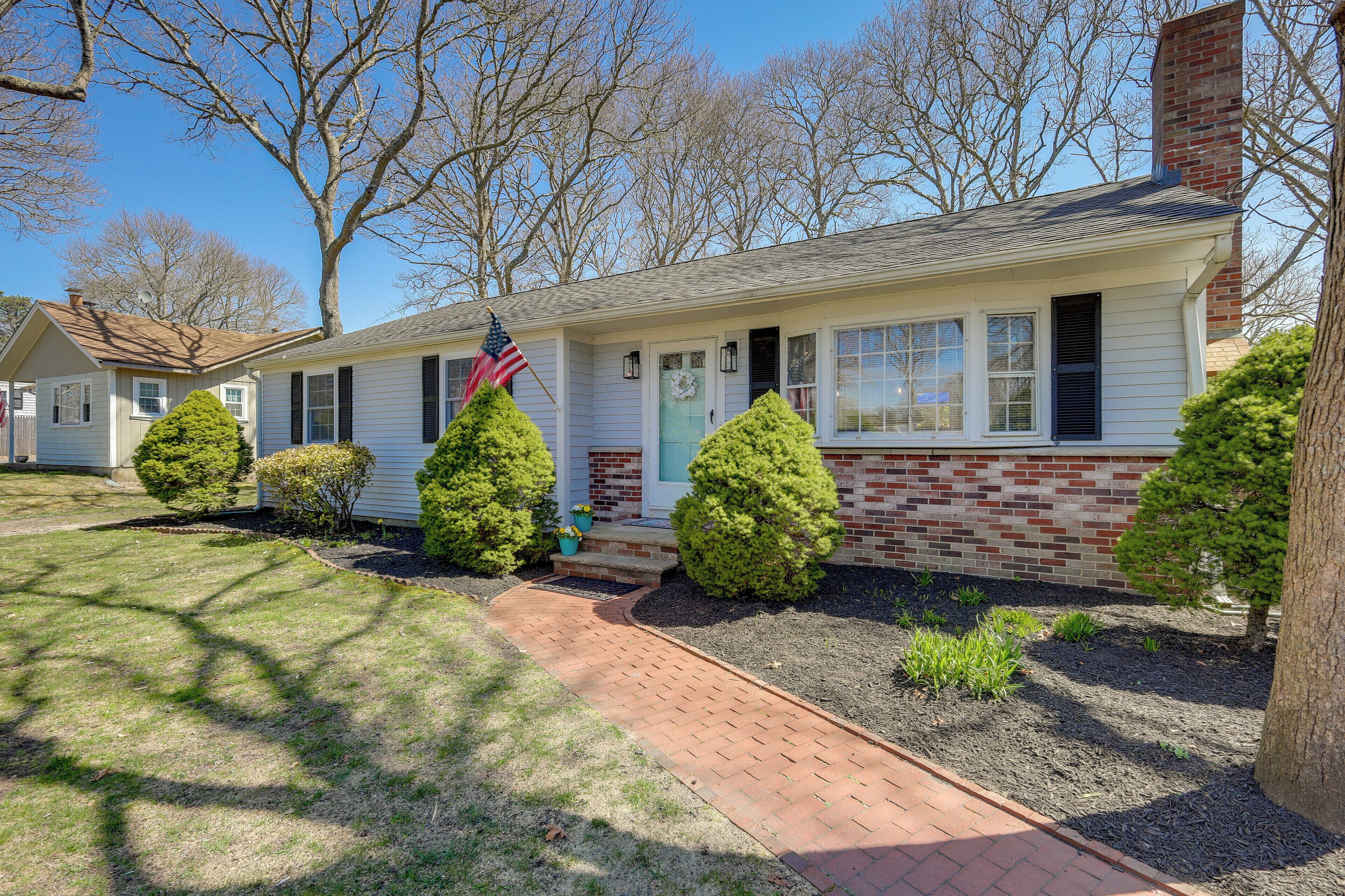 A single-story house with a brick foundation and white siding, a front porch with a small American flag, and a well-maintained lawn with trimmed bushes.