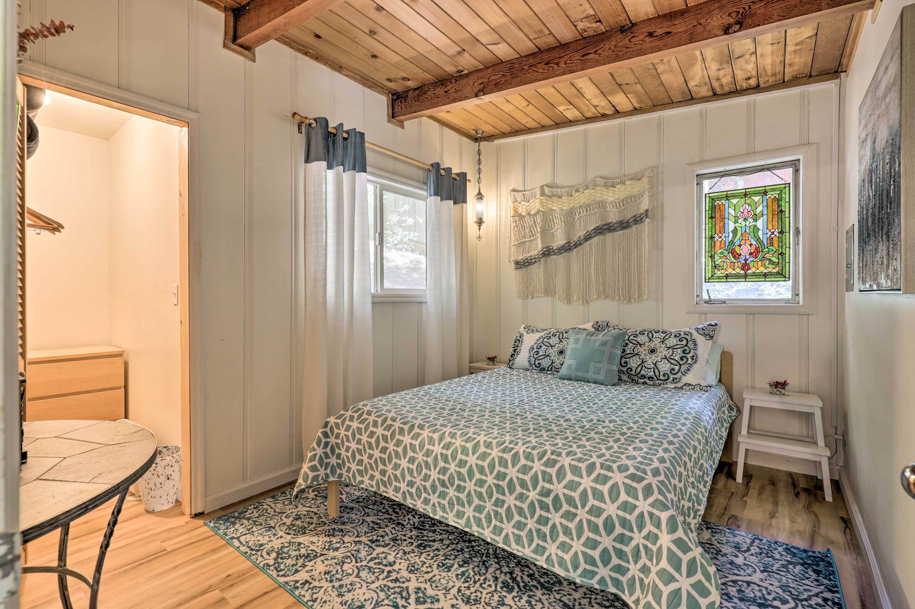 A bedroom with a bed, a small table, a chair, a window with curtains, and a wooden ceiling.
