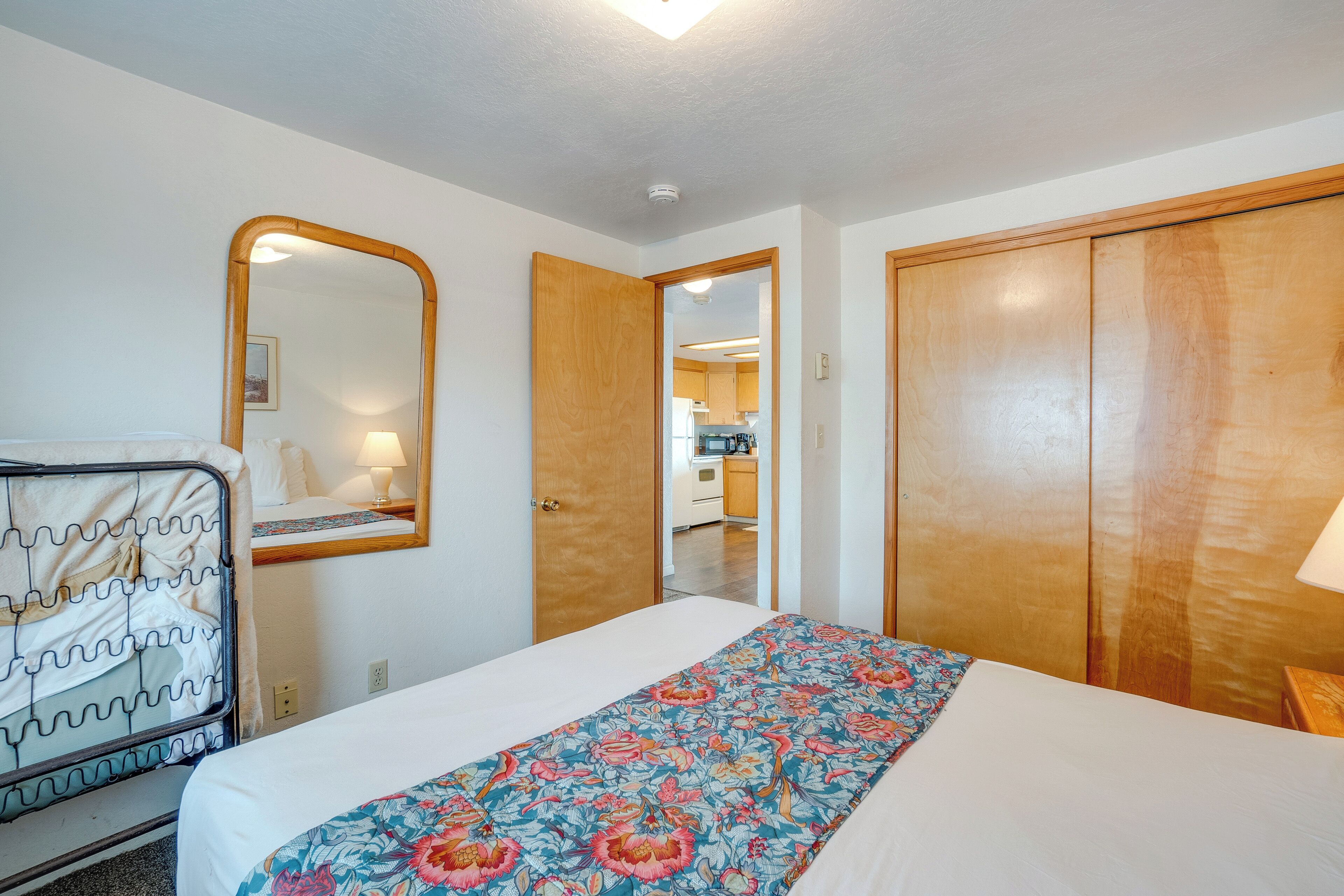 Une chambre à coucher comprenant un lit, une armoire en bois, un miroir et une porte donnant sur la cuisine.