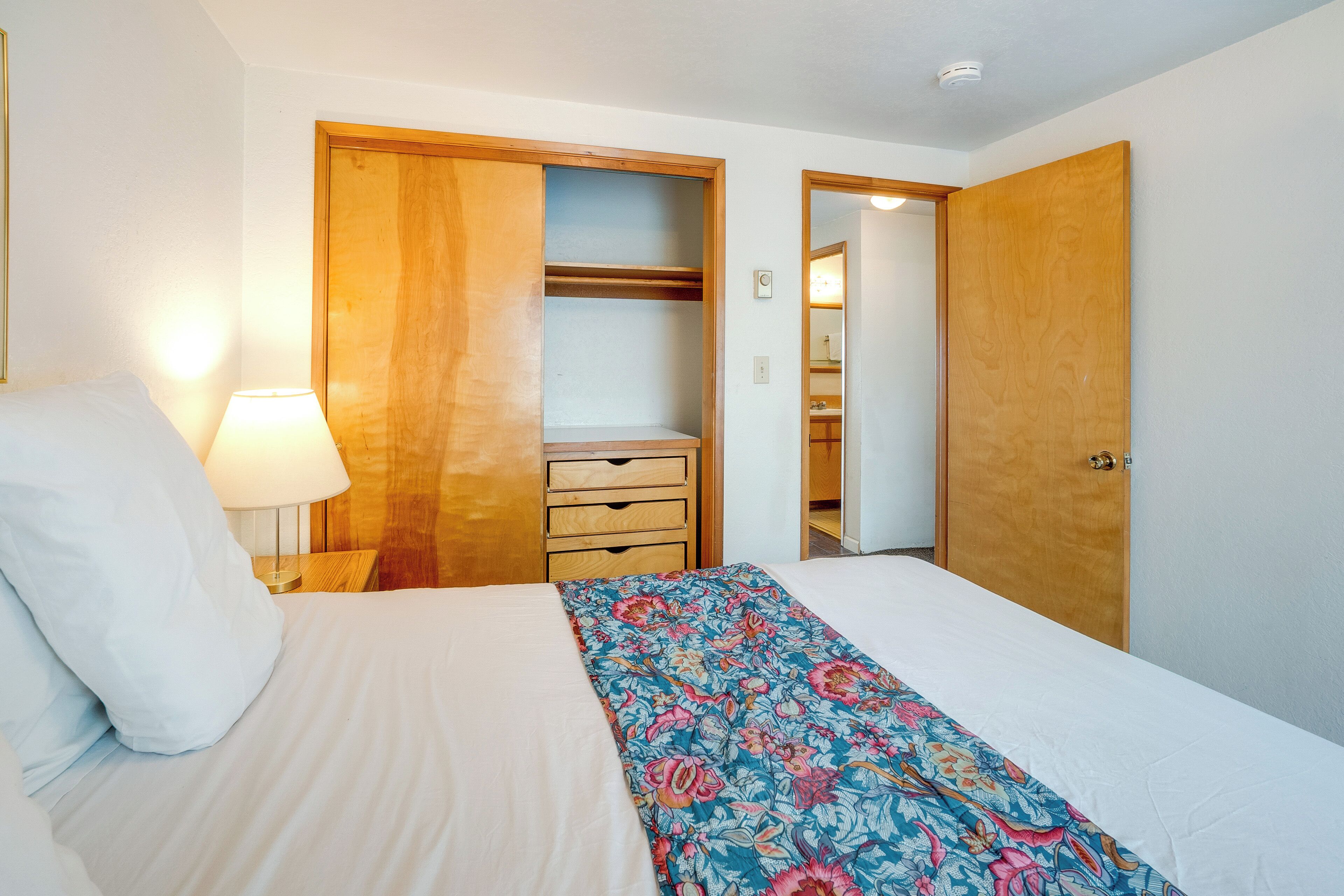 Un dormitorio con cama, mesita de noche, lámpara, armario de madera y baño visible a través de una puerta abierta.