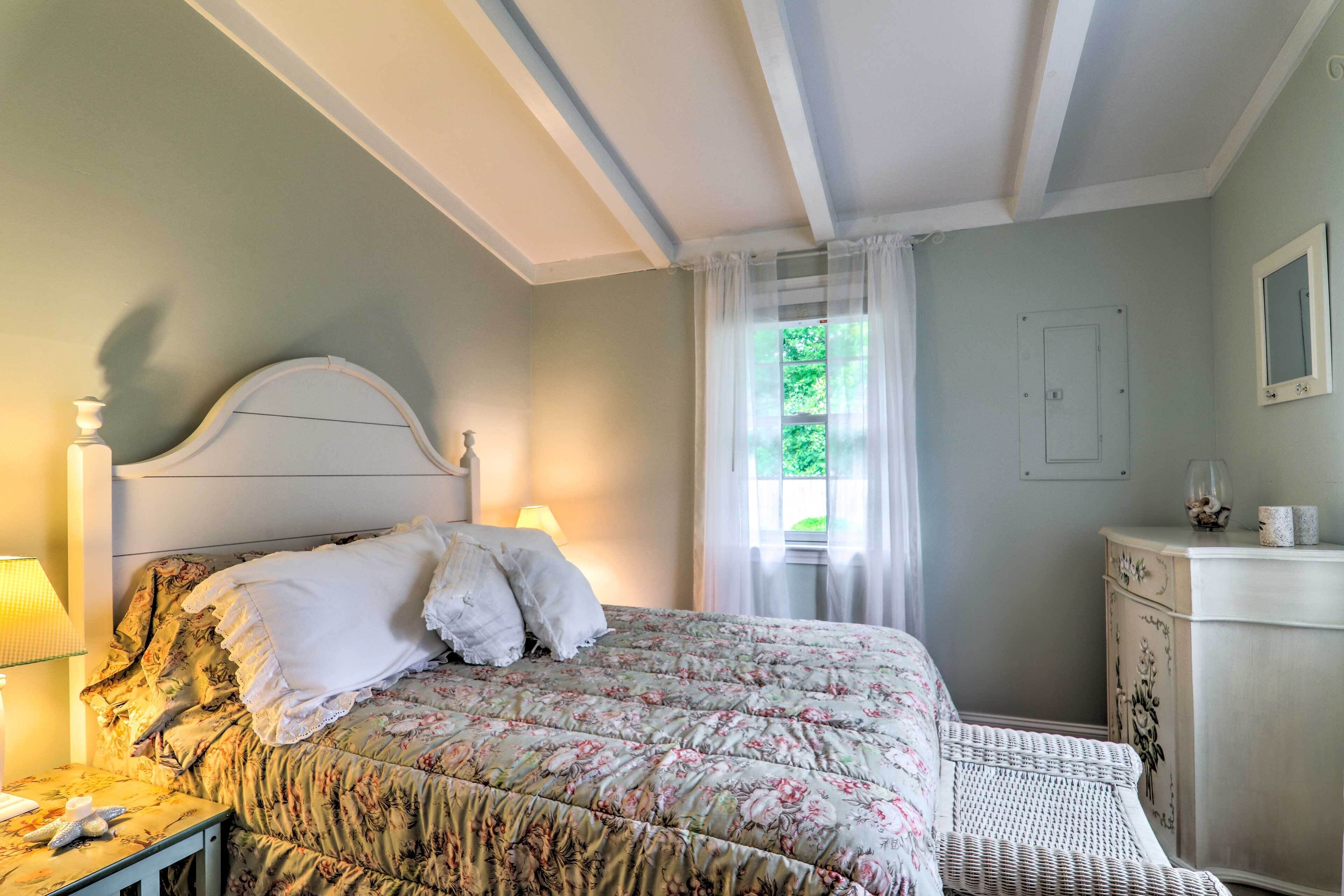 A bedroom with a floral bedspread, a bedside lamp, a dresser, and a window with sheer curtains.