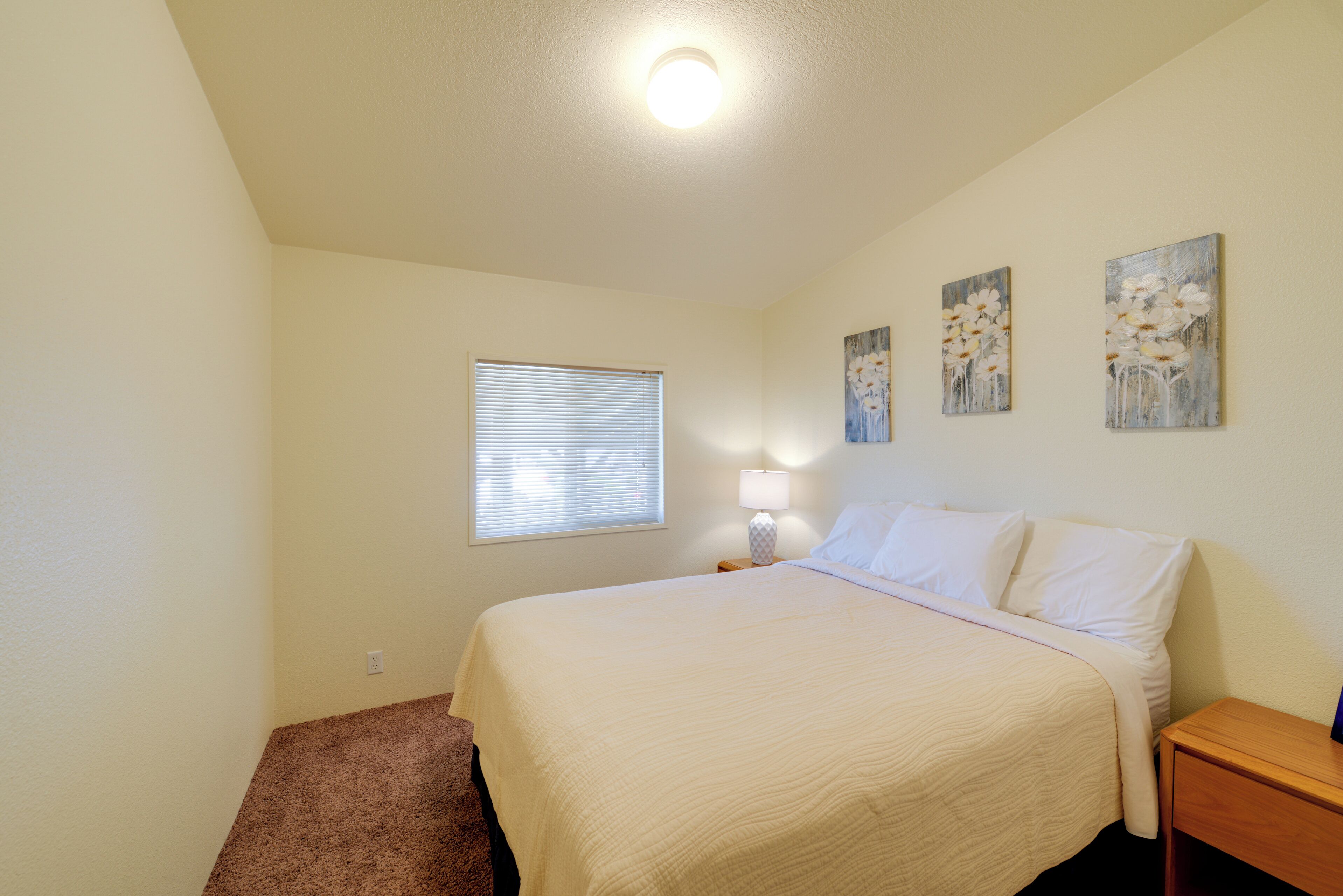 A bedroom with a bed, a nightstand, a lamp, a window with blinds, and two wall-mounted paintings.