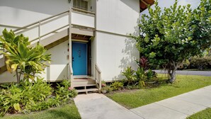 A two-story building with a blue door, a concrete pathway leading to the entrance, and various plants around the property.