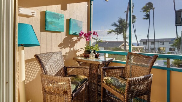 A balcony with wicker chairs, a table with a vase of flowers, and a view of palm trees and buildings.